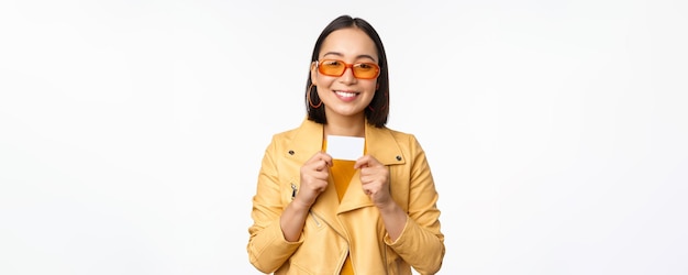Портрет улыбающейся корейской модели в солнцезащитных очках с кредитной картой, стоящей на белом фоне