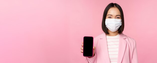 Portrait of smiling korean saleswoman in medical face mask business suit showing smartphone screen standing over pink background