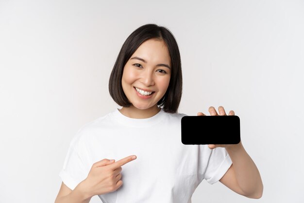 Portrait of smiling korean woman pointing finger at mobile phone screen showing horizontal smartphone display recommending website or store online white background