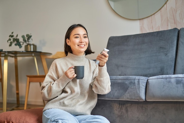 Ritratto di una donna coreana sorridente seduta vicino alla tv che tiene il telecomando e cambia canale mentre beve