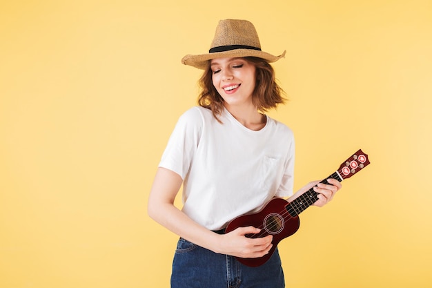 Ritratto di signora sorridente in cappello di paglia in piedi con una piccola chitarra e felicemente giocando su di esso su sfondo rosa