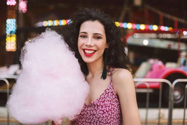 Free photo portrait of smiling lady with dark curly hair in dress standing with cotton candy in hand and happily looking in camera while spending time in amusement park