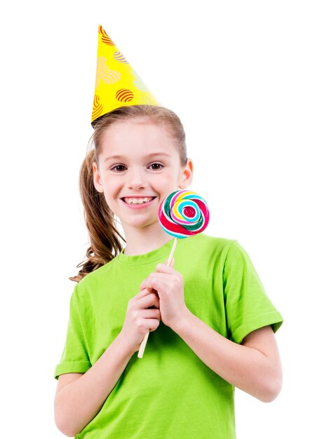 Portrait of smiling little girl in green t-shirt and party hat with colored candy - isolated on white