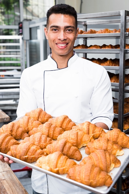 Portrait of a smiling male baker holding tray of baked croissant