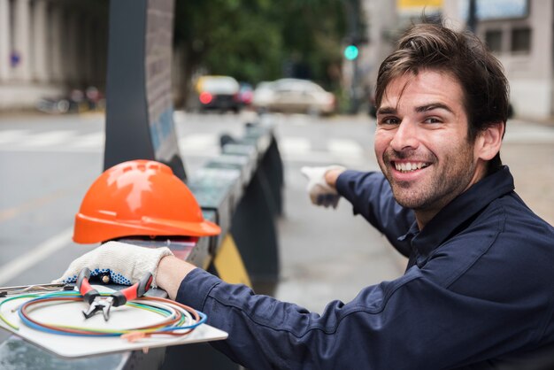 Portrait of smiling male electrician pointing with hard hat and equipment on street