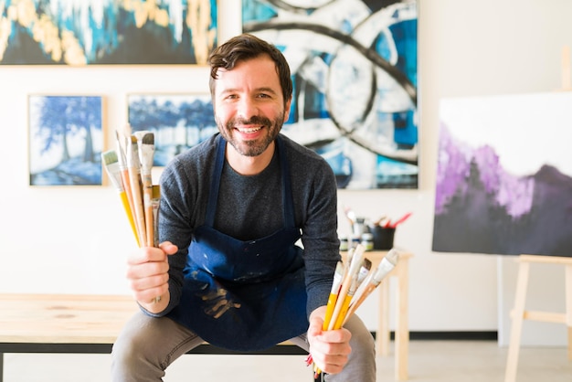 Portrait of a smiling male painter showing his collection of paint brushes and sitting next to a colorful painting in an art gallery