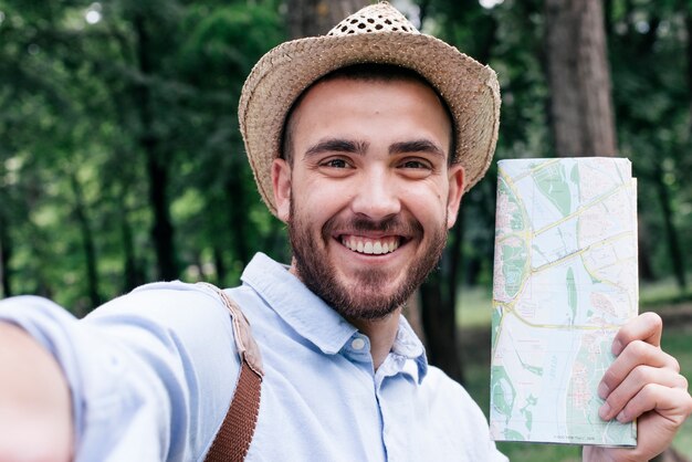 Portrait of smiling man holding map taking selfie at outdoors