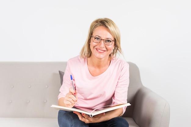 Ritratto di donna matura sorridente con penna e libro seduto sul divano guardando la fotocamera