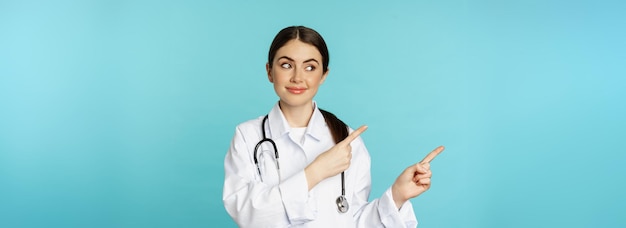 Портрет улыбающегося медицинского работника-врача в белом халате со стетоскопом, указывающим пальцем влево