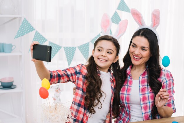 Портрет улыбающейся матери и дочери с ушками зайчика на голове, делающей селфи на мобильном телефоне