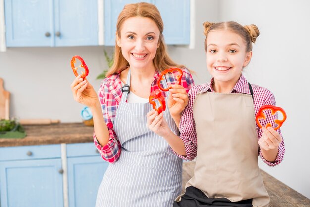 Portrait of smiling mother and her daughter looking at camera holding bell pepper slice in hand