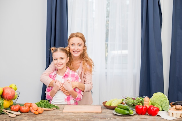 Free photo portrait of a smiling mother and her daughter standing behind the wooden table looking at camera