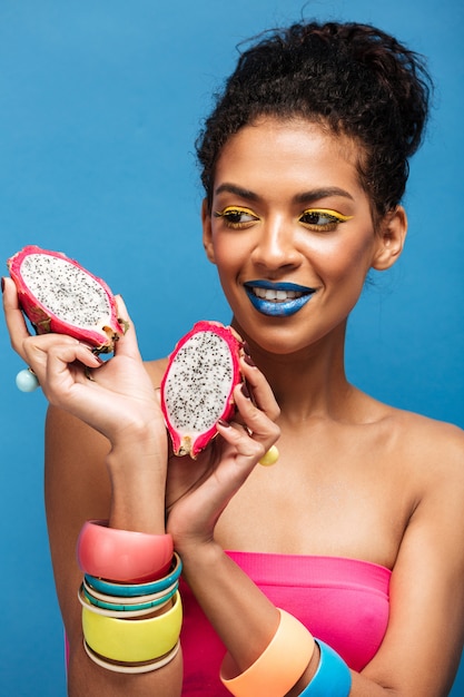 Portrait of smiling mulatto woman with bright cosmetics on face tasting ripe pitahaya cut in half isolated , over blue wall