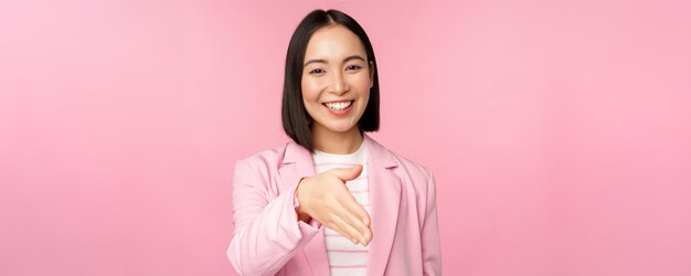 Portrait of smiling pleasant businesswoman shaking hands with business partner handshake extending hand and saying hello standing over pink background
