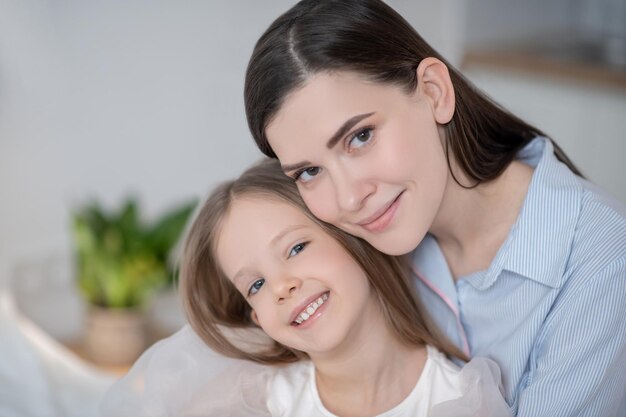 Portrait of a smiling pleased beautiful young dark-haired woman hugging a cheerful adorable little girl