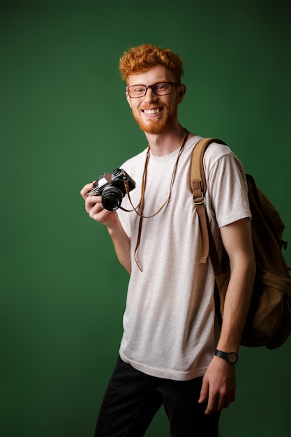 Portrait of smiling readhead bearded hipster with retro camera and backpack