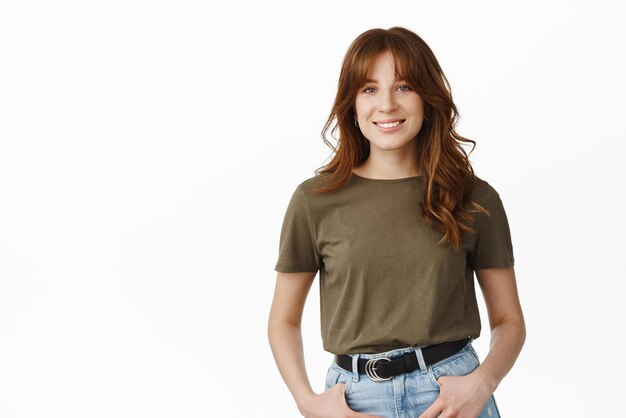 Portrait of smiling redhead girl with bangs looking cute and happy at camera casually standing relaxed pose with hands in jeans pockets white background