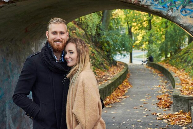 Portrait of smiling redhead male and cute blonde female under old bridge arch in autumn park.