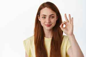 Free photo portrait of smiling redhead woman looks confident shows okay zero sign no proble gesture assure you guarantee quality standing over white background