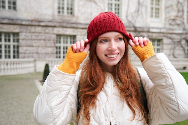 Free photo portrait of smiling redhead woman puts on red hat and smiles wearing warm clothes while exploring ci