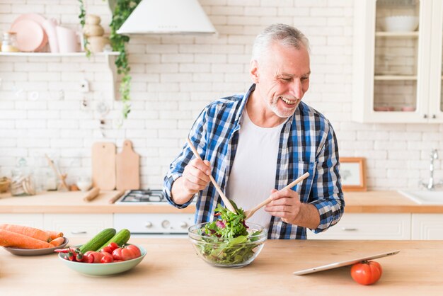Портрет улыбающегося старшего человека, глядя на цифровой планшет, готовит салат на кухне