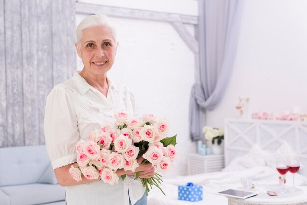 Портрет улыбающейся старшей женщины, держащей букет розовых цветов дома