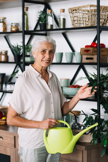 Portrait of smiling senior woman holding watering can touching plant at home