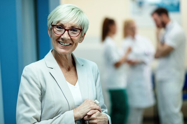 Portrait of smiling senior woman looking at camera while standing in a corridor at the hospital Team of doctors is in the background