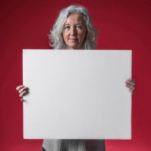 Portrait of a smiling senior woman showing white blank placard against red backdrop