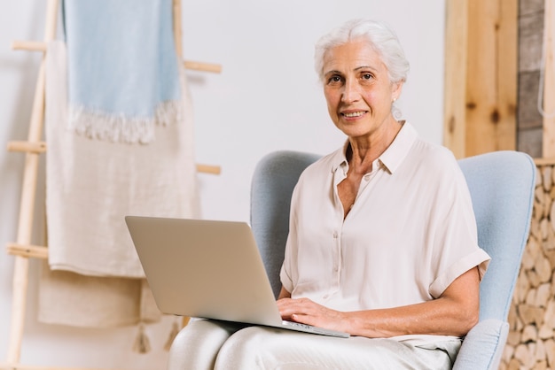 Ritratto della donna senior sorridente che si siede sulla sedia con il computer portatile che esamina macchina fotografica