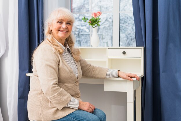 Портрет улыбающейся старшей женщины, сидящей перед окном около стола