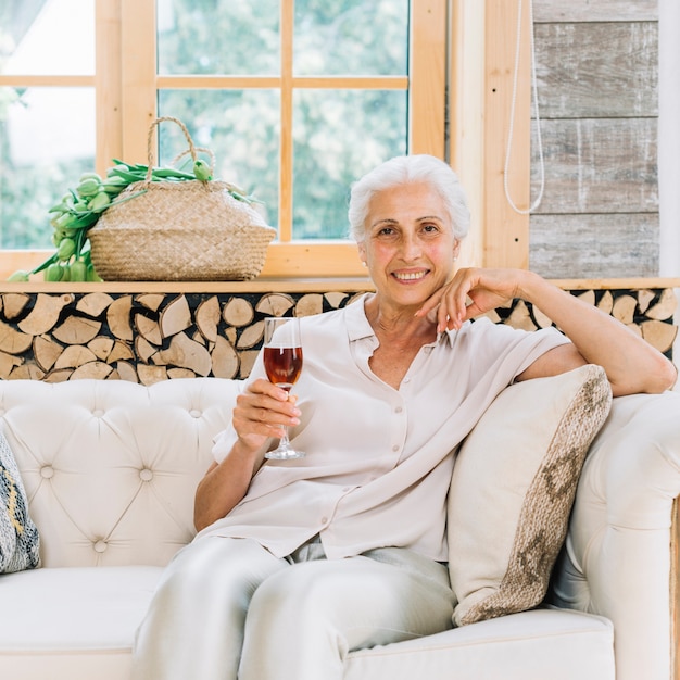 Foto gratuita ritratto della donna senior sorridente che si siede sul sofà che tiene bicchiere di vino