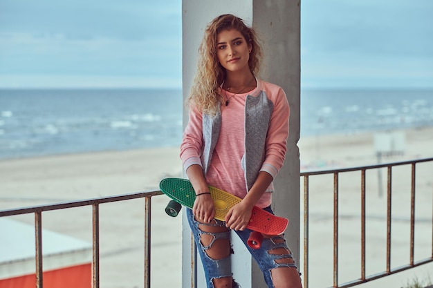 Portrait of a smiling sensual young girl with blonde hair dressed in a pink jacket standing near a guardrail against sea coast.