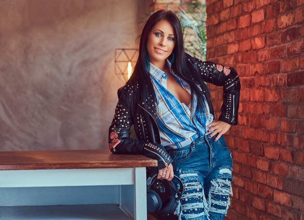 Free photo portrait of a smiling sexy sensual brunette posing in a stylish leather jacket and jeans leaning on a table in a studio with a loft interior.