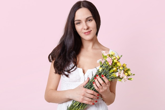 Портрет улыбающегося искреннего молодого модель, стоящего над светло-розовым, держа в руках красочные весенние цветы