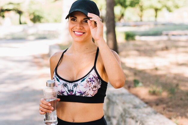 Портрет улыбающегося спортивной молодой женщины в черной кепке держит пластиковую бутылку с водой