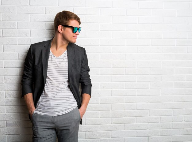 Портрет улыбающегося стильного человека в солнцезащитных очках, стоящего у кирпичной стены в современном офисе.