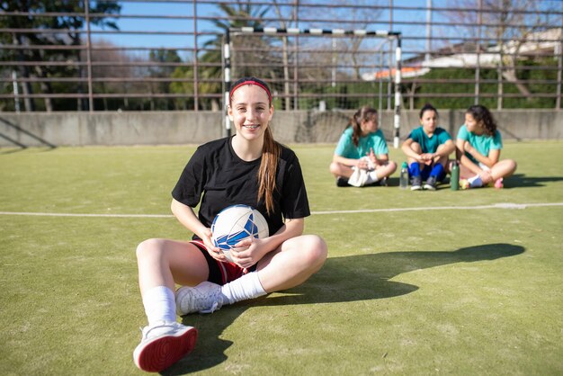 フィールドで笑顔の10代のサッカーの女の子の肖像画。他の女の子が彼女の後ろに座って話を楽しんでいる間、カメラを見て地面に休んでいるスポーツウェアの幸せな女の子。アクティブな休息とチームスポーツのコンセプト