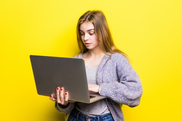 黄色の壁に分離されたラップトップコンピューターを保持している笑顔の10代の少女の肖像画