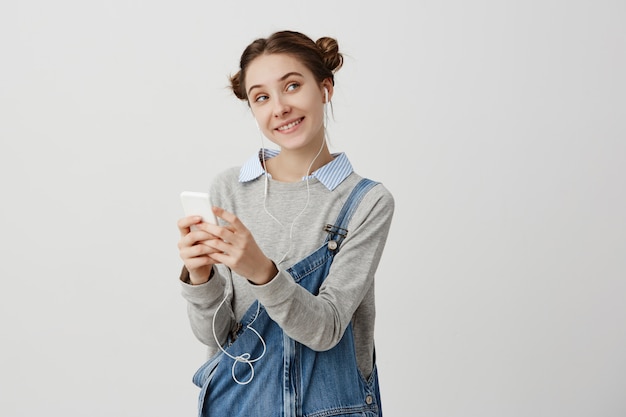 Portrait of smiling woman in denim jumpsuit looking sideways with tricky eyes. with headphones. Funny female office worker chatting with her colleague being flirty. Positive feelings