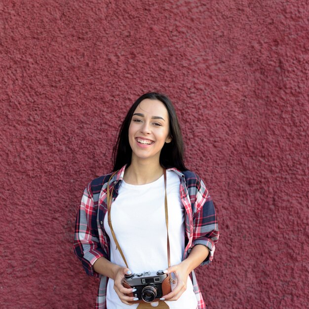 Portrait of smiling woman holding camera standing against maroon wall
