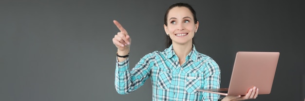 Portrait of smiling woman holding laptop and pointing finger to side choosing destination for