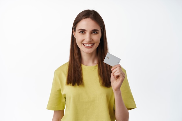 Portrait of smiling woman paying with credit card, advertisement of contactless payment, money transfer or cashback system, standing against white background. Online shopping.