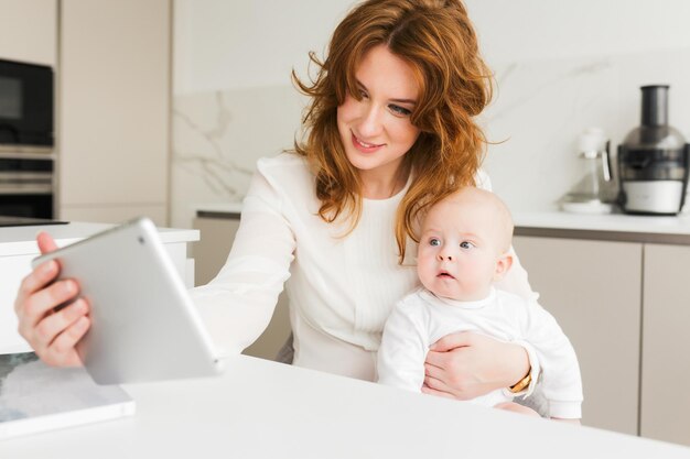 태블릿을 행복하게 사용하면서 귀여운 아기를 손에 안고 앉아 웃고 있는 여성의 초상화