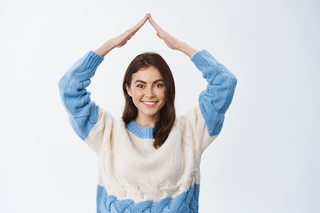 Portrait of smiling woman in sweater showing home roof, making rooftop hands and looking joyful at front, white wall