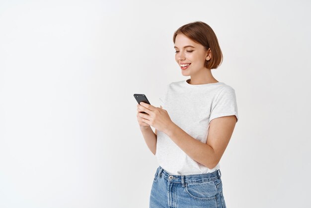 전화 화면을 읽고 웃는 여자 문자 메시지의 초상화 행복 한 여자는 흰색 배경에 서 있는 앱에서 채팅