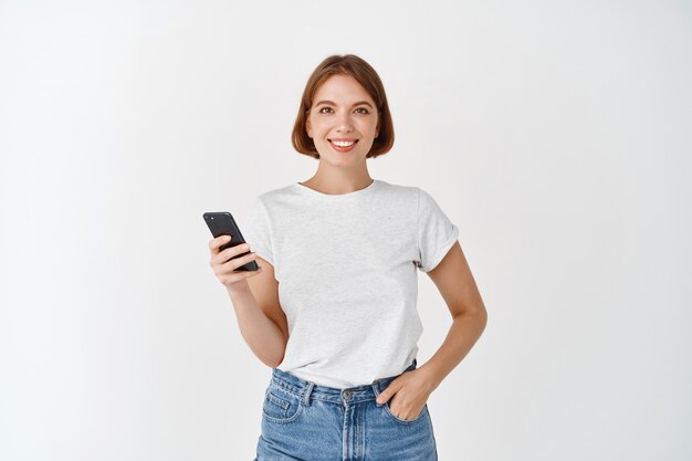 Portrait of smiling woman using smartphone, chatting on social media, standing with cell phone against white wall