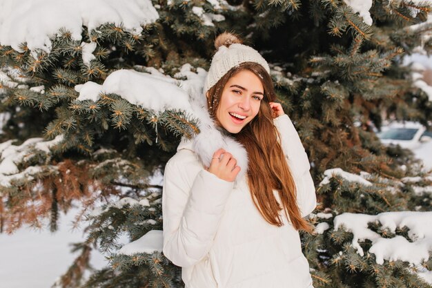 Portrait of smiling woman in warm white coat posing beside tree in frosty day. Outdoor photo of romantic lady with long hair standing in front of snowy spruce during winter photoshoot.