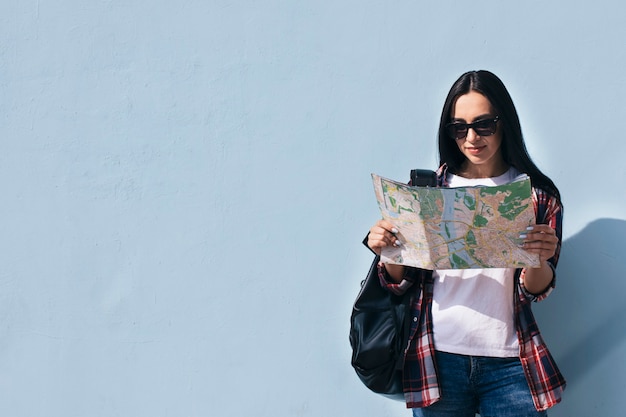 Foto gratuita ritratto degli occhiali da sole d'uso sorridenti della donna che leggono mappa e che stanno vicino alla parete blu