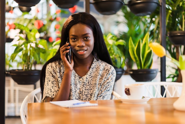 Портрет улыбающейся молодой африканской женщины, сидящей в кафе, делая телефонный звонок
